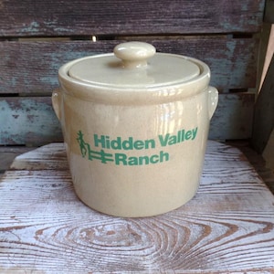May include: Tan ceramic crock with a lid and two handles. The crock has the words "Hidden Valley Ranch" in green lettering. The crock is sitting on a wooden surface.