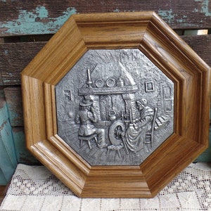 May include: A framed pewter wall plaque depicting a scene of two people working in a workshop. The plaque is in a dark wood octagonal frame.
