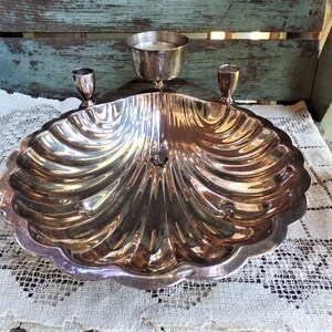May include: A silver-plated seashell-shaped dish with two candle holders. The dish is designed to hold small items, such as snacks or jewelry.