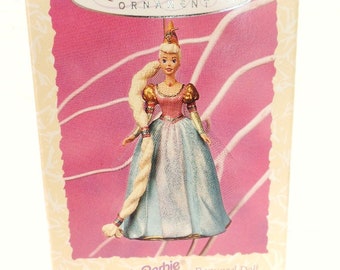 May include: A white box with a pink background featuring a Barbie doll dressed as Rapunzel. The box reads "Keepsake Ornament" and "Based on the Barbie as Rapunzel Doll Children's Collector Series New Collector's Series".