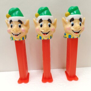May include: Three red and white Pez dispensers with elf heads. The elves are wearing green hats with white trim and yellow and blue striped scarves.