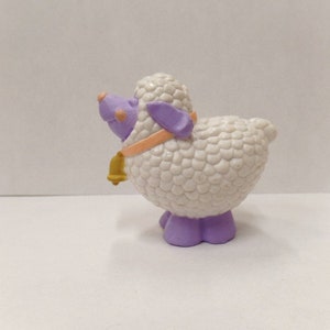May include: A white plastic toy sheep with a purple head and legs. The sheep has a bell around its neck and a pink collar.