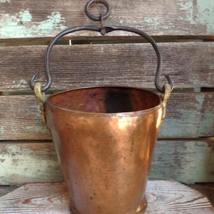 May include: A copper bucket with a dark metal handle and a hanging ring. The bucket has a rounded base and a slightly flared top. The handle is attached to the bucket with rope. The bucket is set against a weathered wooden background.