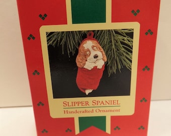 May include: A red and white handcrafted ornament of a sleeping slipper spaniel dog. The ornament is packaged in a red box with a green ribbon and the words "SLIPPER SPANIEL Handcrafted Ornament".