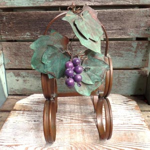 May include: A decorative metal grape vine sculpture with copper-colored frame and leaves. The leaves are green with copper accents, and the grapes are purple. The sculpture is designed to stand upright and is suitable for indoor or outdoor display.