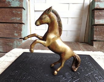 May include: A gold-colored metal horse figurine standing on its hind legs with its front legs raised. The horse has a detailed mane and tail. The figurine is on a black surface with a decorative pattern.