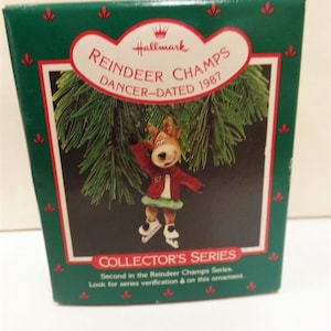 May include: A green and white box with a Hallmark Reindeer Champs ornament inside. The ornament is a reindeer wearing a red jacket and green skirt, ice skating. The box says "Reindeer Champs Dancer-Dated 1987" and "Collector's Series".
