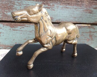 May include: A gold-colored metal horse figurine with a detailed design. The horse is standing on its hind legs with its front legs raised in a running pose.