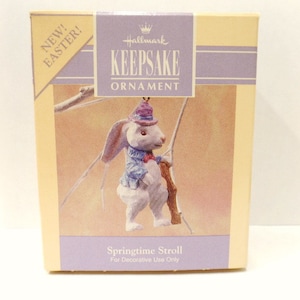 May include: A white rabbit wearing a blue and purple outfit and a hat is holding a stick. The rabbit is a Hallmark Keepsake Ornament. The box reads "New! Easter!" and "Springtime Stroll".