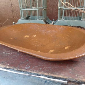 May include: A large, brown wooden dough bowl with a slightly oval shape. The bowl has a smooth, worn surface with a light brown patina.