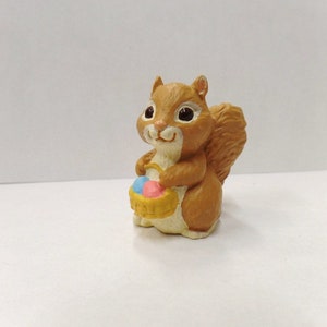 May include: A brown squirrel figurine holding a basket of colorful Easter eggs. The squirrel has a bushy tail and is standing on a white surface.