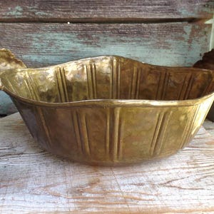 May include: A vintage brass bowl with a hammered texture and vertical ribbed design. The bowl has decorative grape cluster handles and sits on a weathered wooden surface. The bowl's color is a warm, antique gold.
