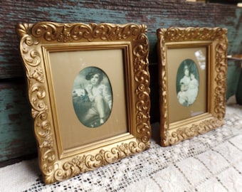 May include: Two ornate gold picture frames with detailed carvings. The frames are on a white lace tablecloth. The frames contain black and white portraits.