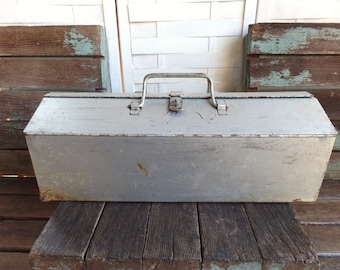 May include: A vintage metal toolbox with a silver finish and a carrying handle. The toolbox has a worn, distressed look and is sitting on a wooden surface.