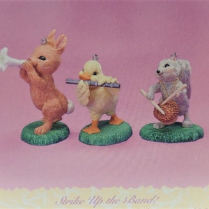 Puede incluir: Tres peque&ntilde;as figuritas de un conejo tocando una trompeta, un pato tocando una flauta y una ardilla tocando un tambor. Las figuritas est&aacute;n sobre un fondo rosa con el texto "Strike Up the Band!" en la parte inferior.