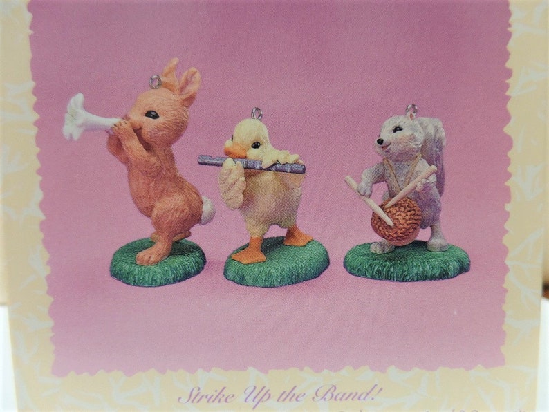 Puede incluir: Tres peque&ntilde;as figuritas de un conejo tocando una trompeta, un pato tocando una flauta y una ardilla tocando un tambor. Las figuritas est&aacute;n sobre un fondo rosa con el texto "Strike Up the Band!" en la parte inferior.