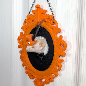 May include: An orange framed wall hanging featuring a white mouse wearing a miniature orange top hat and black bow tie. The mouse is set against a black background within an oval frame. A black chain and decorative orange frame complete the piece.