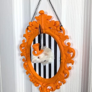 May include: An orange, ornate picture frame with a black and white striped background. Inside the frame is a white mouse wearing an orange top hat with a skull emblem. A black feather is attached to the frame. The frame hangs from a black chain.
