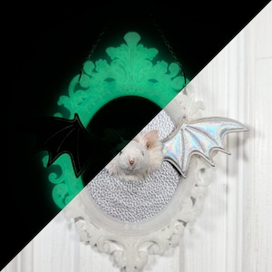 May include: An oval wall hanging featuring a white mouse head centered on a textured silver background within an ornate white frame. The frame glows green in the dark, and the mouse has iridescent, holographic bat wings attached to its sides.