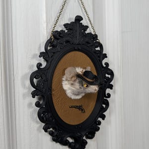 May include: An oval wall hanging featuring a taxidermy mouse head wearing a small black cowboy hat, mounted on a tan textured background within an ornate black frame. A miniature metal revolver is attached below the mouse, and a chain is attached.