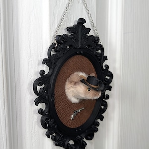 May include: A taxidermy mouse head wearing a small black cowboy hat, mounted on a brown textured oval plaque within an ornate black frame. A miniature silver-colored revolver is attached below the mouse head. The frame hangs from a silver chain.