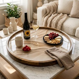 Könnte beinhalten: Ein runder Serviertablett aus Holz mit einer transparenten, wellenförmigen Oberfläche enthält eine Weinflasche, ein Weinglas, einen Teller mit Crackern, Käse, Trauben und eine kleine Schale mit Himbeeren. Eine gefaltete Leinen-Serviette liegt auf dem Tablett.