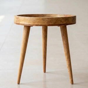 May include: A round side table constructed from light brown wood with a visible grain pattern. The table features a circular top with a raised edge and is supported by three tapered legs that extend outwards from the centre to provide stability.