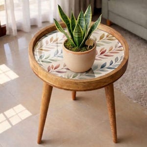 May include: A round wooden side table with three legs. The tabletop features a colorful leaf pattern and holds a potted snake plant. The plant pot is a light brown color. The table is made of light brown wood.