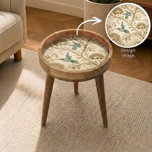 May include: A round wooden side table with three legs. The tabletop features a floral design with hummingbirds in shades of blue, green, and yellow on a cream background. The table is approximately 18 inches in diameter.