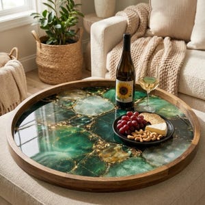 May include: A circular wooden serving tray featuring a green and gold agate design. The tray holds a wine bottle, a glass of wine, a plate of grapes, crackers, cashews, and cheese. The tray is placed on a neutral-toned ottoman.