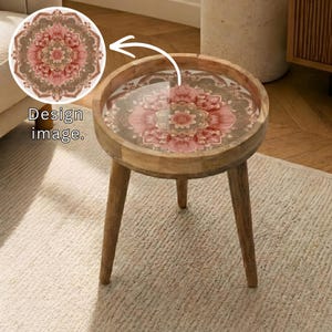 May include: A round wooden side table with a glass top featuring a floral design in shades of pink, brown, and beige. The table has three angled legs and the text "Design image."