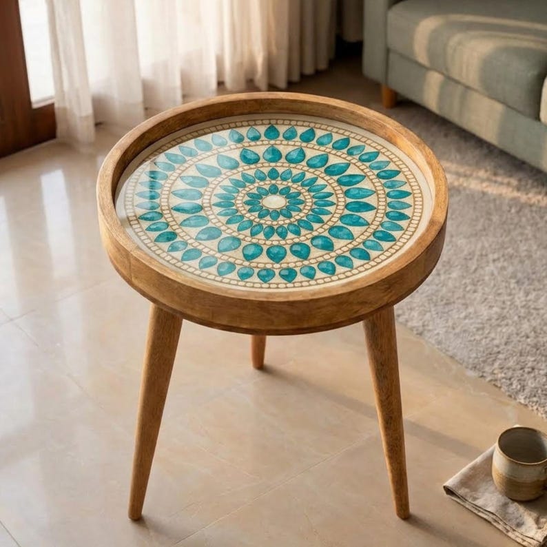 May include: A round wooden side table with three tapered legs and a circular tabletop featuring a decorative inlay. The inlay displays a concentric pattern of teal, teardrop-shaped elements arranged in rings against a cream-coloured background.