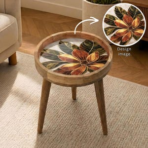 May include: A round wooden side table with a floral design on the tabletop. The design features a large flower with brown, yellow, and green petals. The table has three wooden legs and a light-colored rug beneath it.