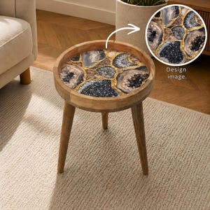 May include: A small, round wooden side table with a decorative top. The tabletop features a geode design with black, gray, and brown sections, accented by gold-colored resin. The table has three angled legs and is set on a light-colored rug.