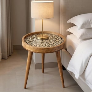 Pu&ograve; includere: Un tavolino rotondo in legno con tre gambe affusolate e un piano circolare caratterizzato da un motivo geometrico intarsiato in bianco e nero. Una lampada in metallo color ottone con paralume cilindrico in tessuto beige &egrave; posata sopra.