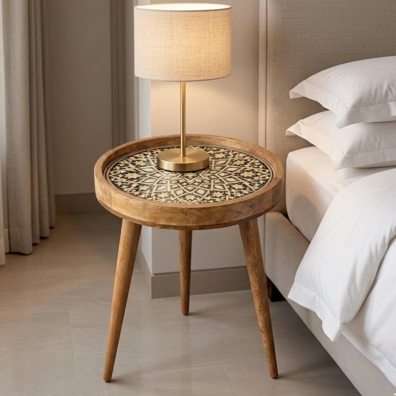 Pu&ograve; includere: Un tavolino rotondo in legno con tre gambe affusolate e un piano circolare caratterizzato da un motivo geometrico intarsiato in bianco e nero. Una lampada in metallo color ottone con paralume cilindrico in tessuto beige &egrave; posata sopra.