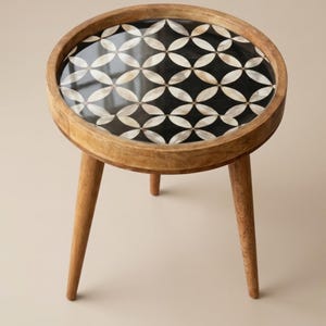 Pode incluir: Uma pequena mesa lateral redonda de madeira com um tampo decorativo. O tampo da mesa apresenta um fundo preto com um padrão repetido de formas de pétalas brancas e creme. Possui três pernas de madeira anguladas.