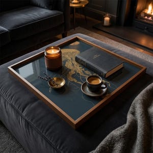 May include: A square wooden tray with a dark blue surface featuring a gold jellyfish design. The tray holds a lit candle, a book, a small bowl, and a cup of coffee. The tray is on a dark gray ottoman.
