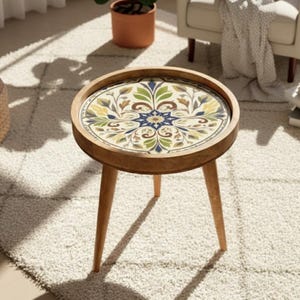 Może przedstawiać: Okrągły drewniany stolik pomocniczy z dekoracyjnym blatem z płytek kwiatowych. Stół ma trzy skośne nogi i podwyższoną krawędź. Płytka ma symetryczny wzór z niebieskimi, zielonymi, żółtymi i brązowymi elementami. Stół ma średnicę około 46 cm.