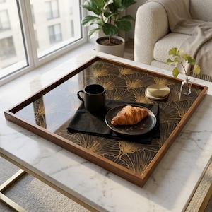 Könnte beinhalten: Ein quadratisches Serviertablett aus Holz mit einem gold-schwarzen Fächermuster. Das Tablett enthält einen schwarzen Becher, eine schwarze Serviette, einen Teller mit einem Croissant und einen kleinen goldfarbenen Behälter. Das Tablett steht auf einem weißen Marmor-Couchtisch.