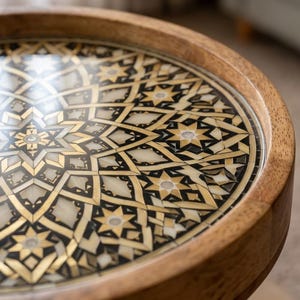 Pu&ograve; includere: Un tavolo rotondo in legno con un intricato design intarsiato sulla superficie. Il piano del tavolo presenta un motivo geometrico composto da metallo dorato, accenti neri e segmenti in pietra o conchiglia color crema a forma di stella.