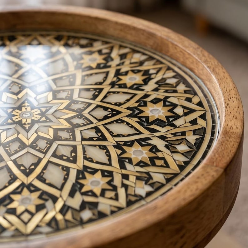 Pu&ograve; includere: Un tavolo rotondo in legno con un intricato design intarsiato sulla superficie. Il piano del tavolo presenta un motivo geometrico composto da metallo dorato, accenti neri e segmenti in pietra o conchiglia color crema a forma di stella.