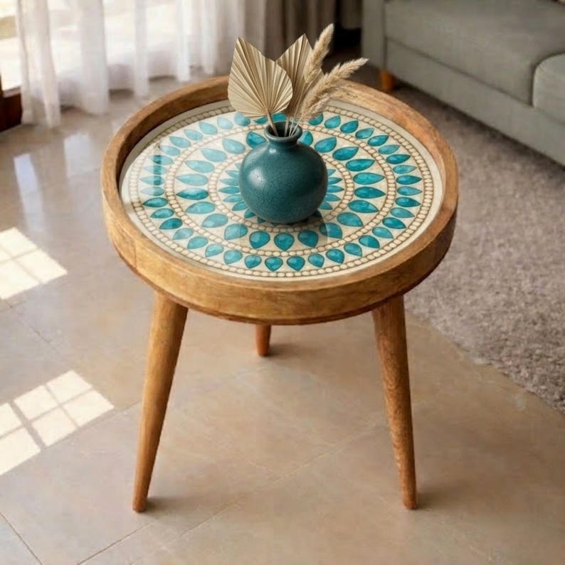 May include: A round wooden side table with three tapered legs and a circular tabletop featuring a decorative inlay of teal and white teardrop patterns. A small, dark teal ceramic vase sits in the centre, holding two dried palm leaves.