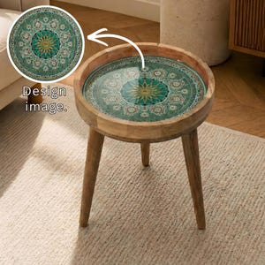 Puede incluir: Una mesa auxiliar redonda de madera con una tapa decorativa estampada en verde azulado, dorado y blanco. La mesa tiene tres patas anguladas y un acabado de madera natural. El texto "Design image" está en la imagen.