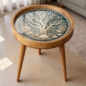 May include: A round wooden side table with a decorative glass top. The glass top features a detailed design of a tree and seashells in white and beige against a teal background. The table has three angled legs.