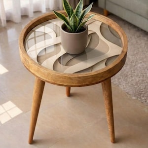 May include: A round wooden side table with three tapered legs and a glass top featuring a layered, wavy beige and grey pattern. A small, grey ceramic pot containing a green snake plant sits centered on the table surface.