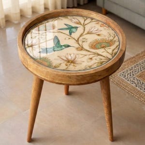 Könnte beinhalten: Ein runder Beistelltisch aus Holz mit dekorativer Glasplatte. Die Glasplatte zeigt ein Blumen- und Kolibri-Design in Grün-, Blau- und Beigetönen. Der Tisch hat drei abgewinkelte Beine und einen erhöhten Rand. Der Tisch hat einen Durchmesser von etwa 51 cm.