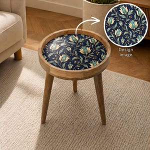 May include: A small, round wooden side table with three legs. The tabletop features a dark blue floral pattern with teal and yellow accents. The design is shown in a close-up view. The table is placed on a light-colored rug.
