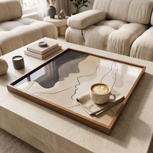 Peut inclure: Un plateau rectangulaire en bois avec un dessus en verre présentant un motif abstrait de deux visages dans des tons gris et beige. Une tasse en céramique tachetée avec un latte et une serviette en lin reposent sur le plateau.