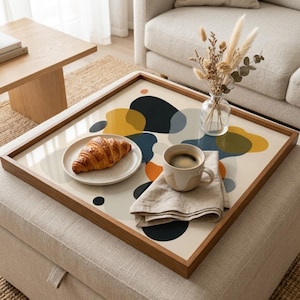 May include: A wooden-framed serving tray with a cream-colored surface featuring abstract shapes in navy, yellow, and orange. A plate with a croissant, a coffee cup, and a linen napkin are on the tray. A vase with dried flowers sits on the tray.